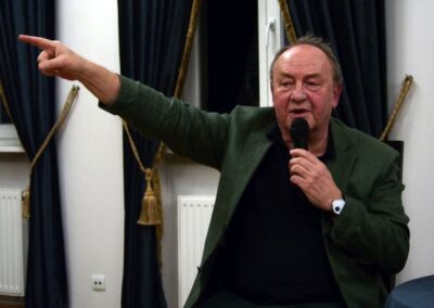 Janusz L. Wiśniewski prawą ręka wskazuje w górę, w drugiej trzyma mikrofon.